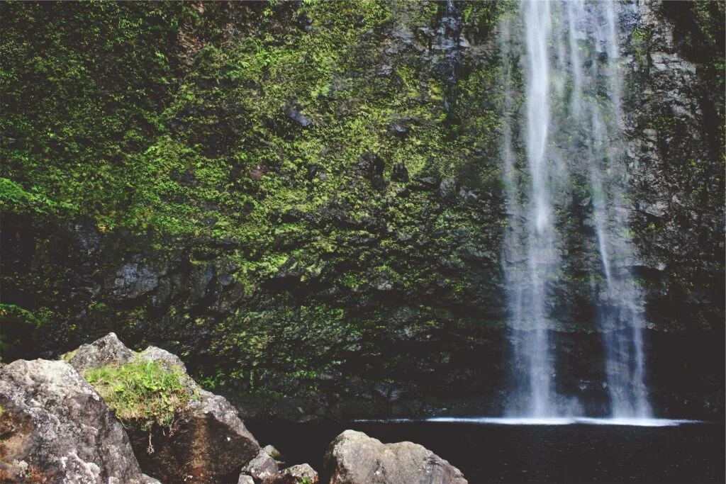 Ein Wasserfall, der über moosbewachsene Felsen in eine dunkle Höhle stürzt.