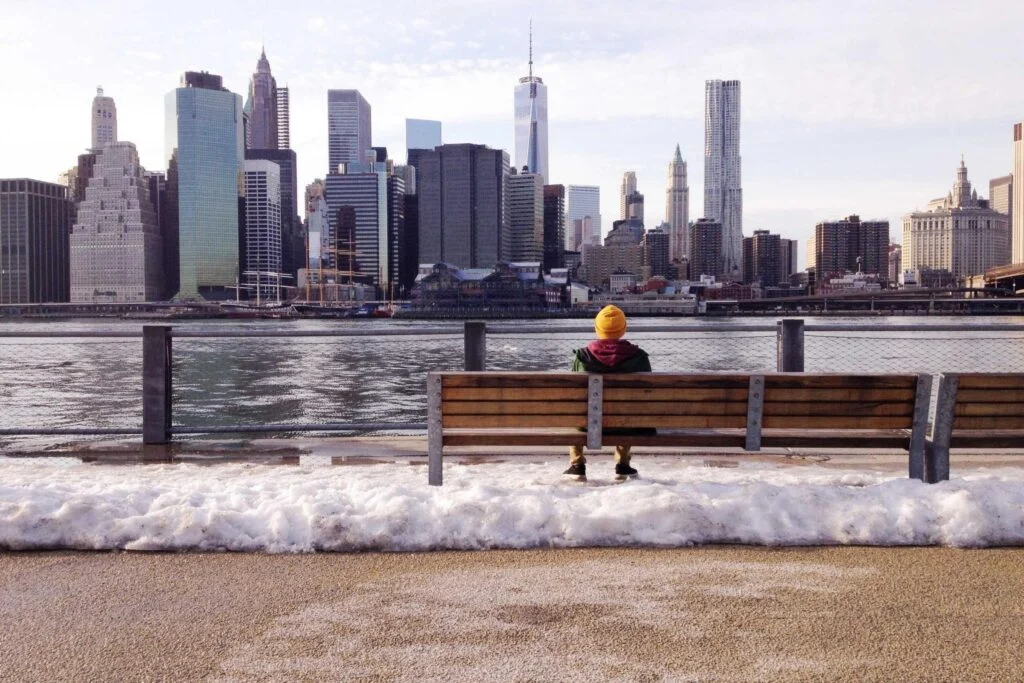 Eine Person sitzt auf einer Bank mit Blick auf eine Stadt mit Wolkenkratzern und Wasser im Hintergrund.